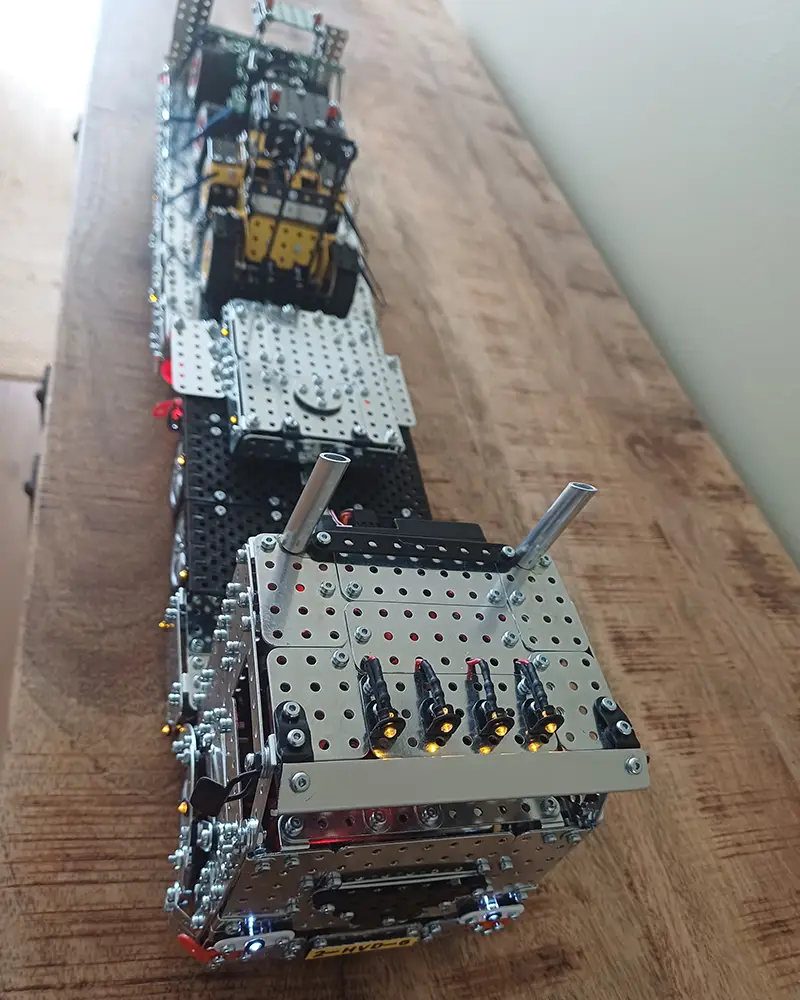 Long metallic construction model on a wooden table, with exposed gears, panels, and small LEDs along the front edge. Designed like a mechanical vehicle or robot built from a metal construction kit.