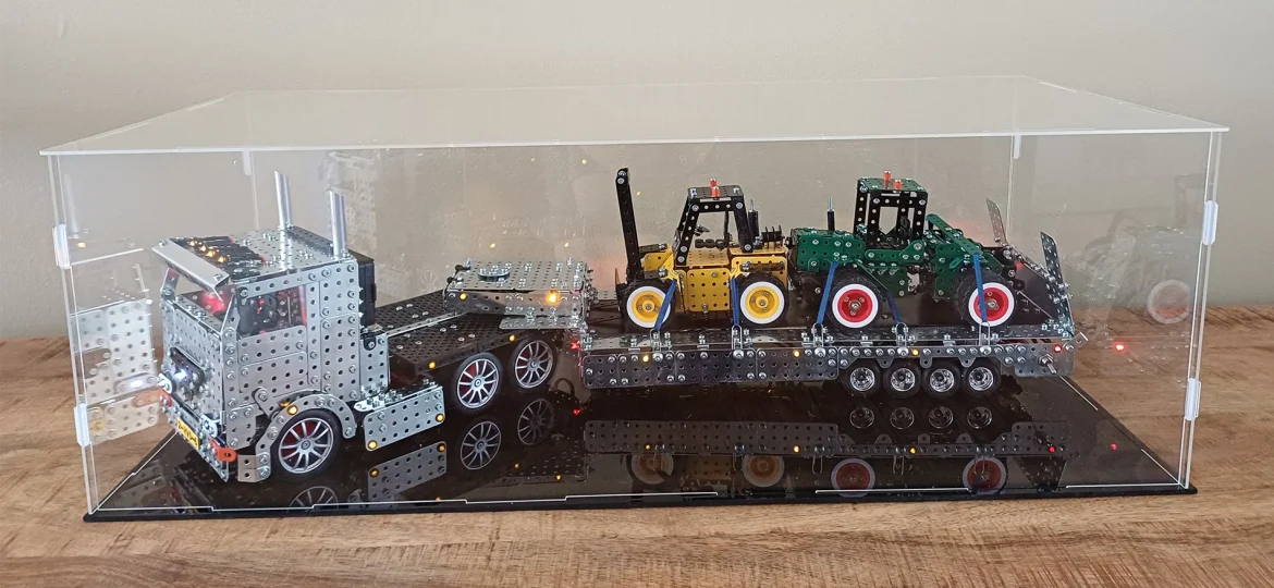 Metal construction toy truck with a long trailer carrying small colored tractor-like models inside a clear display case on a wooden table.