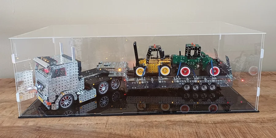 Metal construction toy truck with a long trailer carrying small colored tractor-like models inside a clear display case on a wooden table.